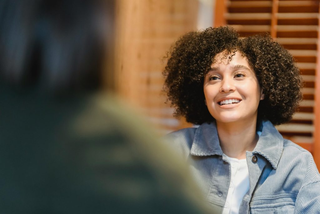 A cheerful young woman with curly hair smiling during a casual indoor conversation.